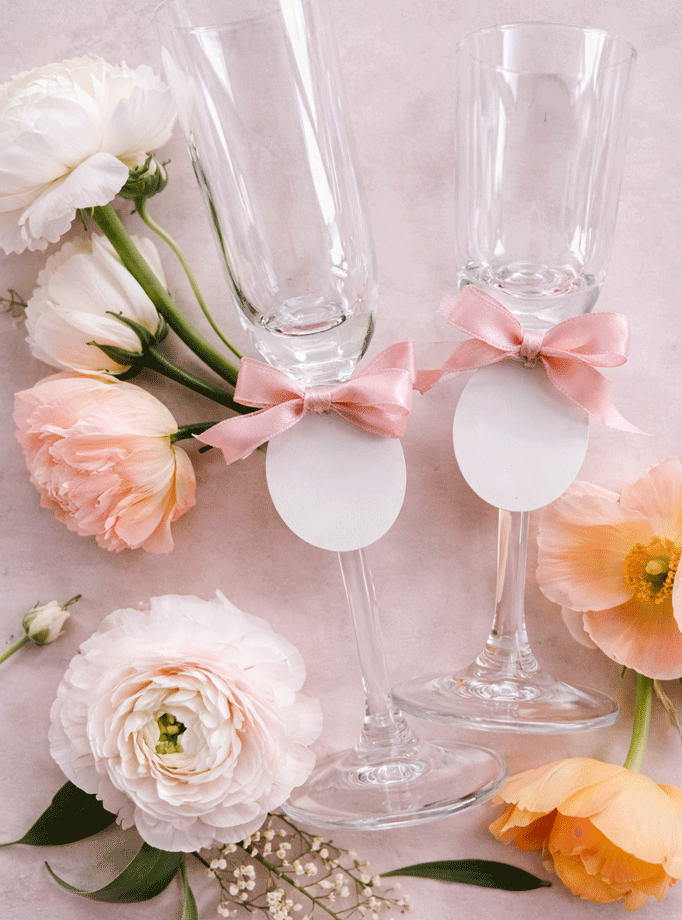 Dvije elegantne kristalne čaše za šampanjac ukrašene ružičastim mašnama i personaliziranim okruglim karticama pored nježnog cvijeća.