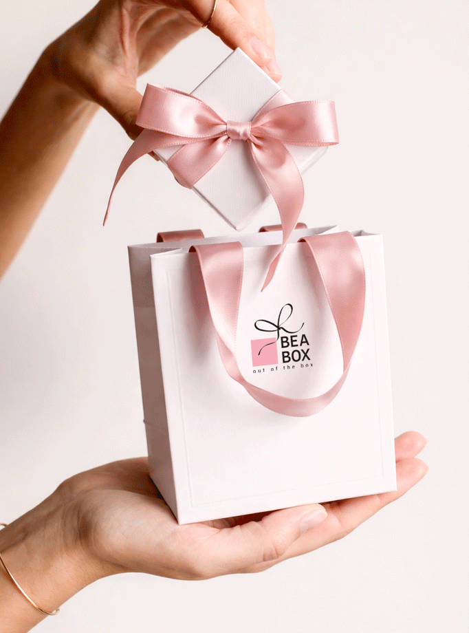 Ženska ruka drži malu bijelu poklon kutijicu s ružičastom satenskom mašnom iznad odgovarajuće bijele vrećice s logotipom.
